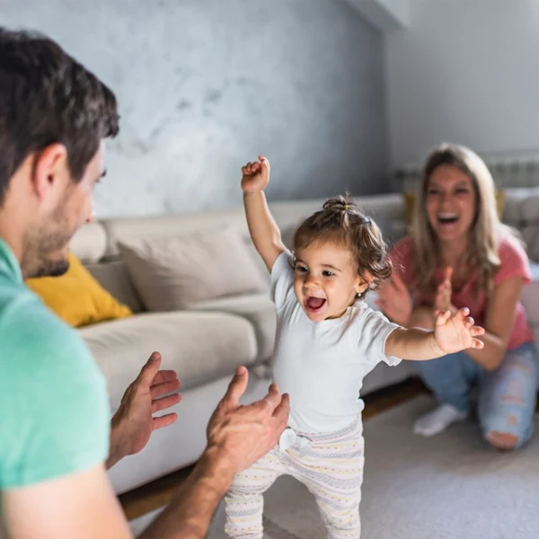 A one year old girl walking from her mom to her dad in their living room