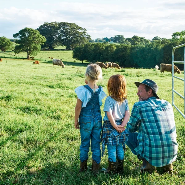 A middle-aged man kneeling next to his two daughters near a gate in a pasture of cows
