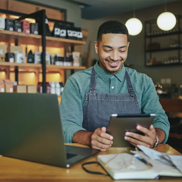 A business owner standing behind a counter at his shop with a laptop and iPad reviewing business documents.